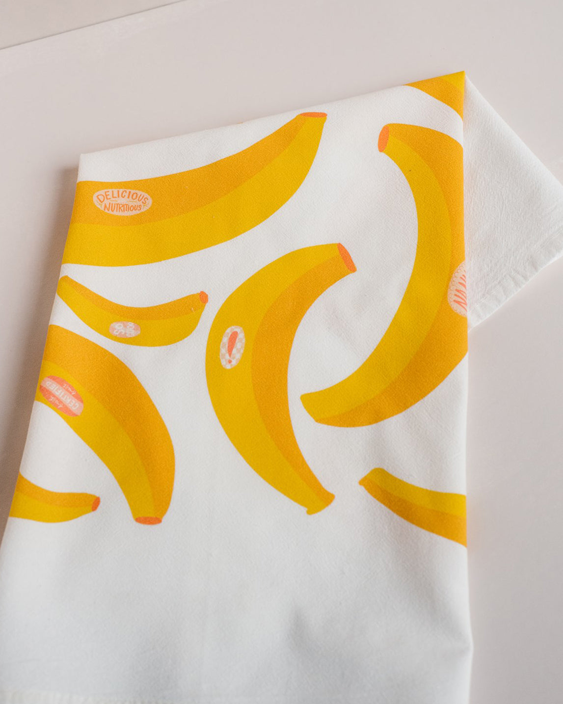 white tea towel with bright yellow bananas with pink fruit stickers printed on it
